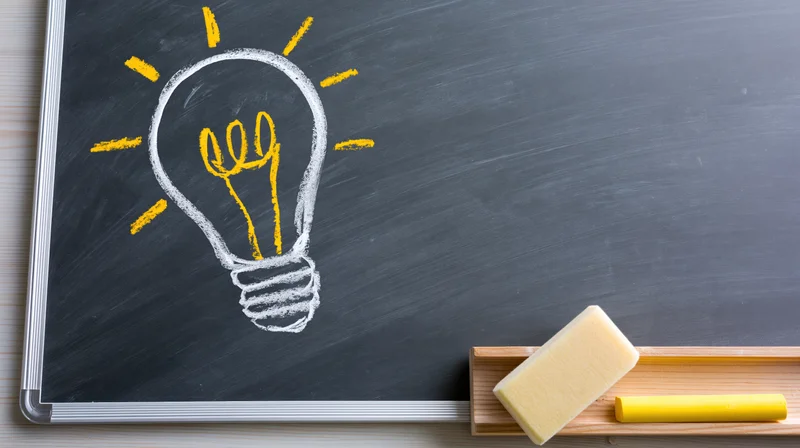 Lightbulb drawn in white and yellow chalk on a blackboard with eraser and chalk in a wooden tray beside it