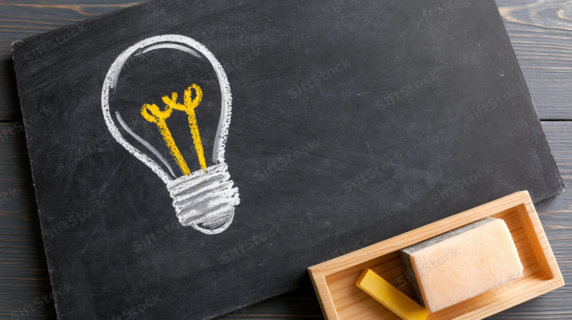 Chalk-drawn lightbulb on blackboard with chalk and eraser in wooden tray on dark wood surface