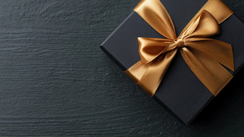 Matte black gift box with gold satin ribbon bow on dark textured surface