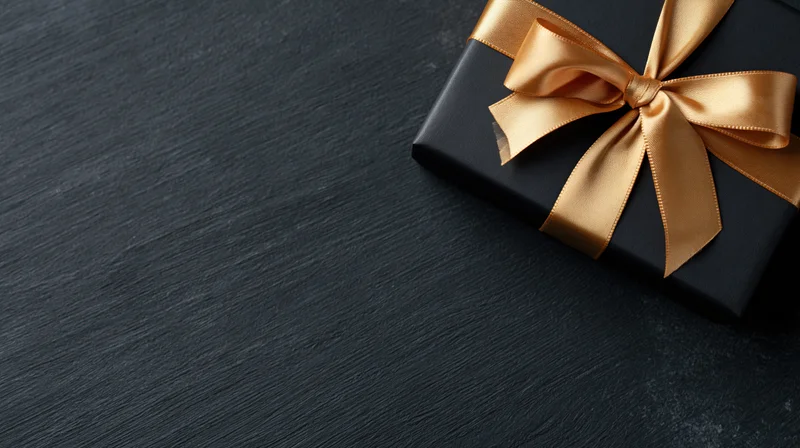 Matte black gift box with gold satin ribbon bow on dark textured surface
