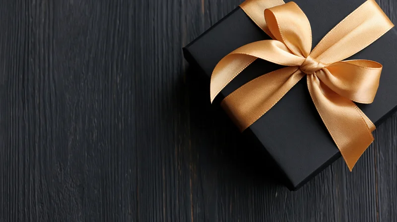Matte black gift box with gold satin ribbon bow on dark wooden surface