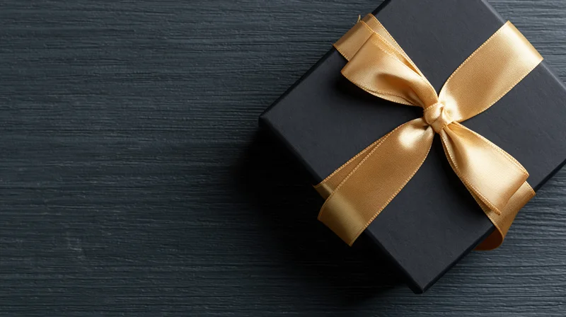 Matte black gift box with gold satin ribbon bow on dark wooden surface