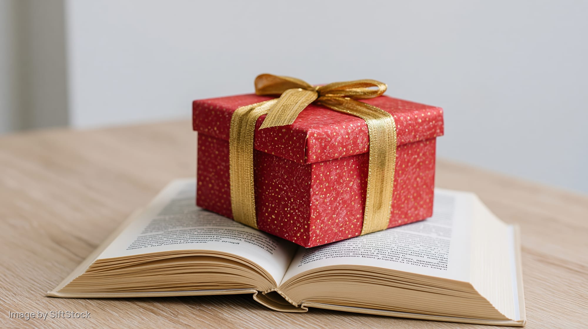 Red gift box with gold ribbon and bow resting on open book on wooden table