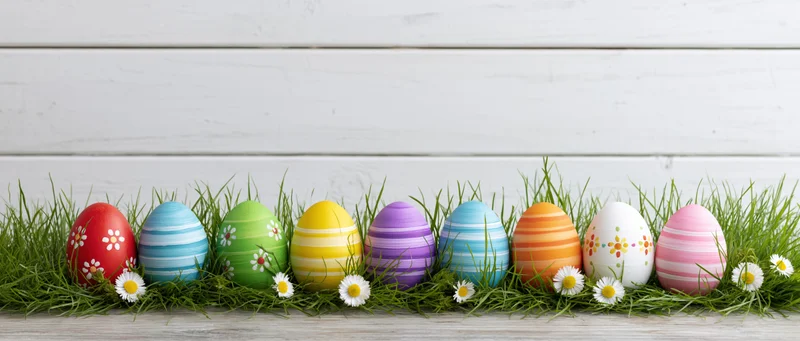 Row of colorful painted easter eggs arranged on grass with daisies against white wooden background