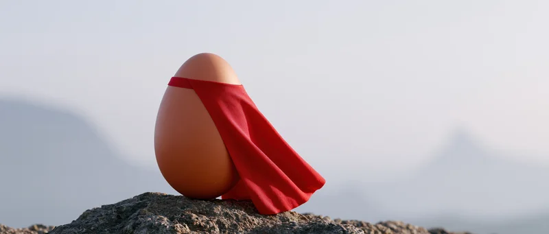 Brown chicken egg with red superhero cape resting on rocky outcrop against misty mountain backdrop