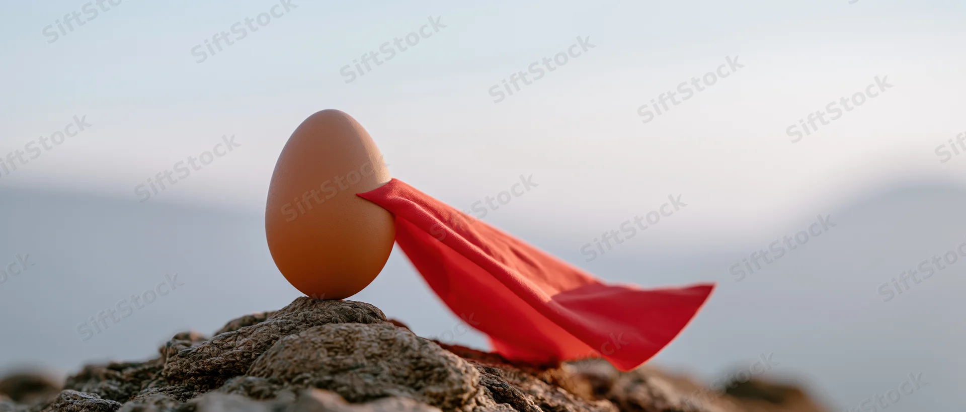 Brown egg with red superhero cape balanced on rocky peak at dawn