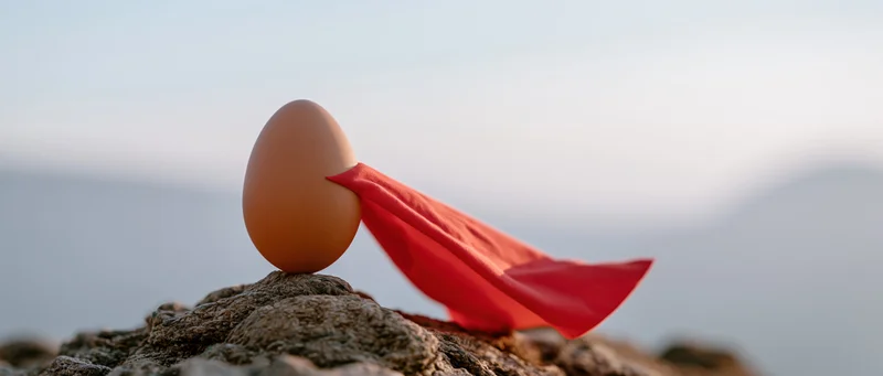Brown egg with red superhero cape balanced on rocky peak at dawn