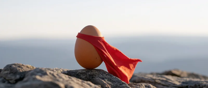 Brown egg with red superhero cape perched on rocky cliff overlooking ocean