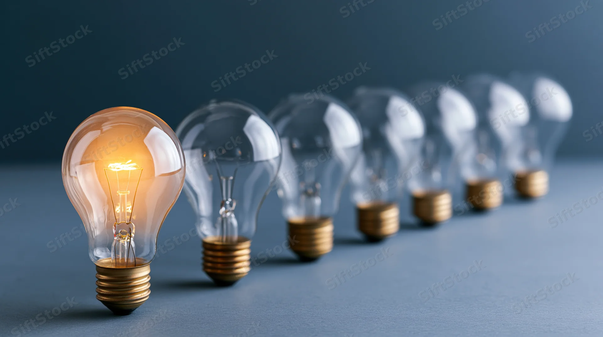 Glowing incandescent lightbulb in front of a row of unlit bulbs on a blue surface