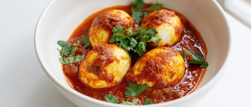 Indian egg curry with halved boiled eggs in red sauce garnished with cilantro in a white ceramic bowl