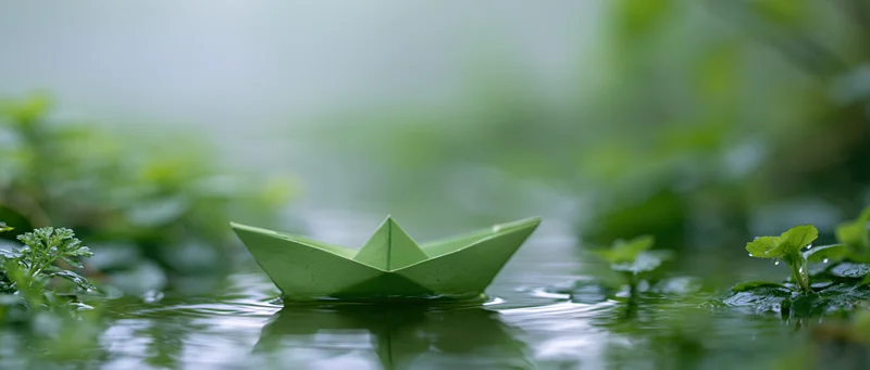 Green paper boat floating on calm shallow water surrounded by foliage