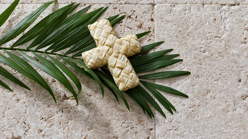 Woven palm frond cross resting on stone tiles with green leaves, flat lay view