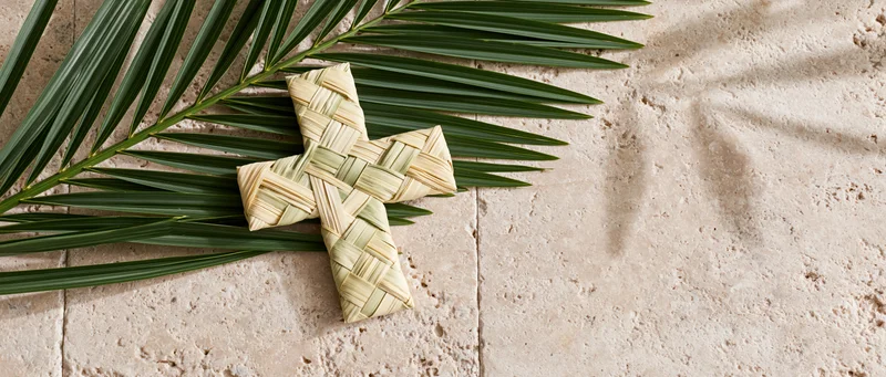 Woven palm frond cross resting on stone tiles with fresh green leaves