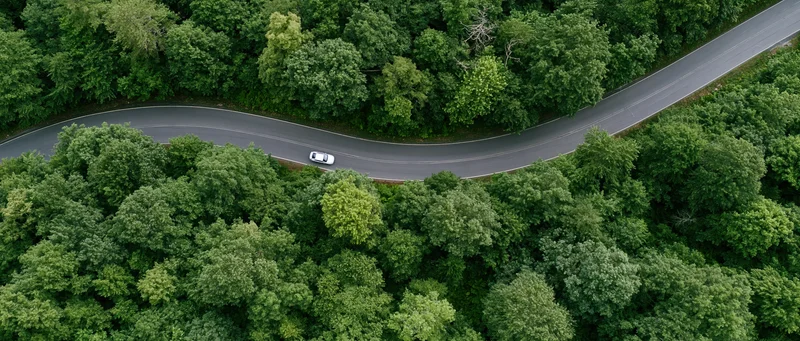 Aerial view of a winding road through dense green forest with a white car driving along it