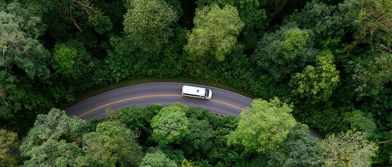 White minibus driving along winding road through dense green forest from aerial top-down view