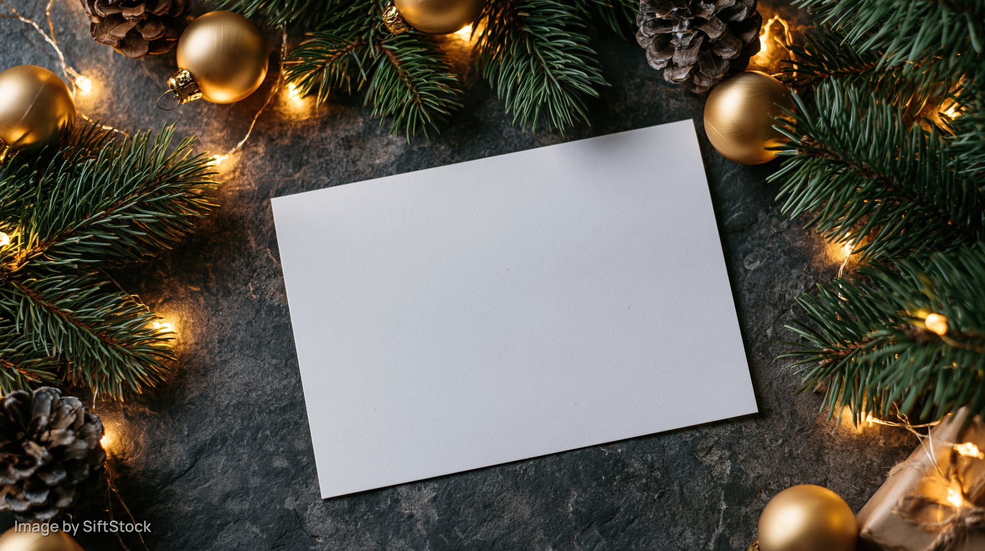 Blank white card on dark stone surface surrounded by christmas decorations and lights