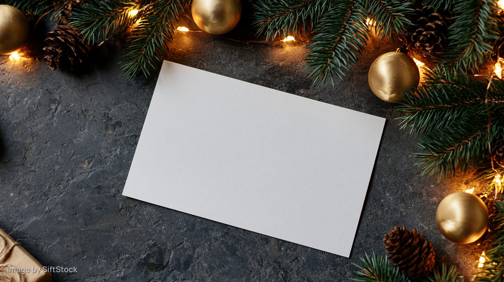 Blank white card on dark stone surface surrounded by christmas garland and gold ornaments