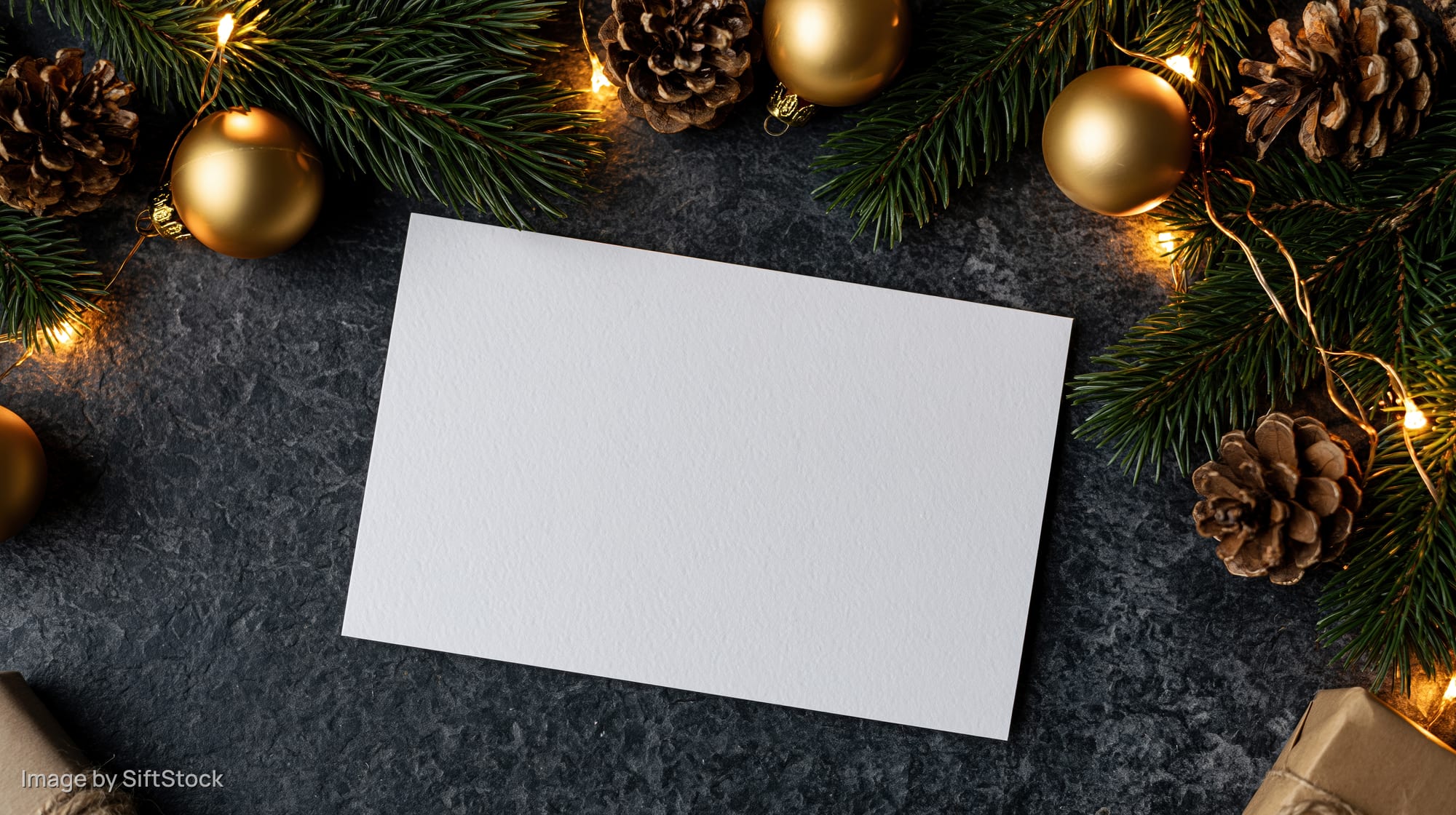 Blank white card on dark stone surface surrounded by pine branches, pinecones, gold ornaments and string lights for christmas decoration