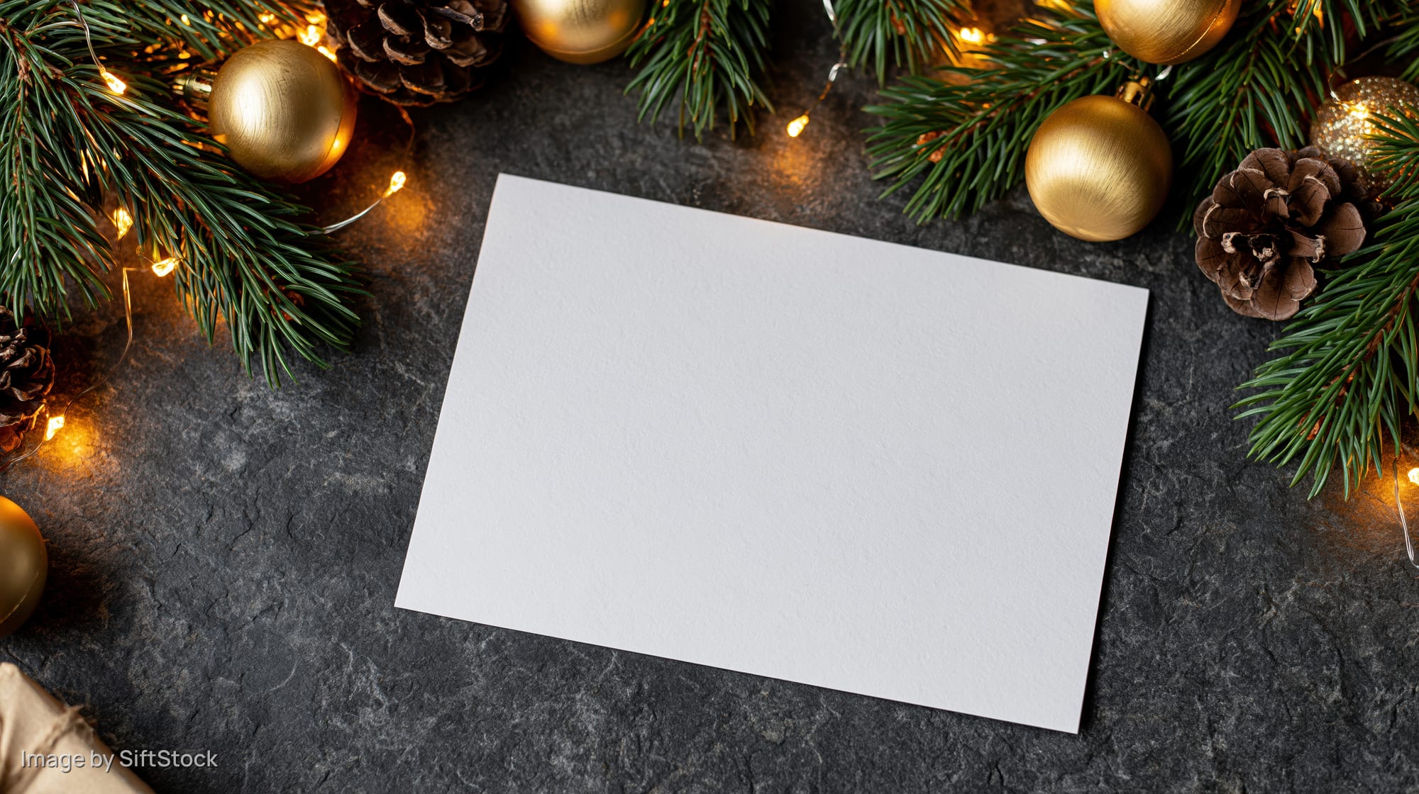 Blank white card on dark stone surface surrounded by christmas decorations and lights