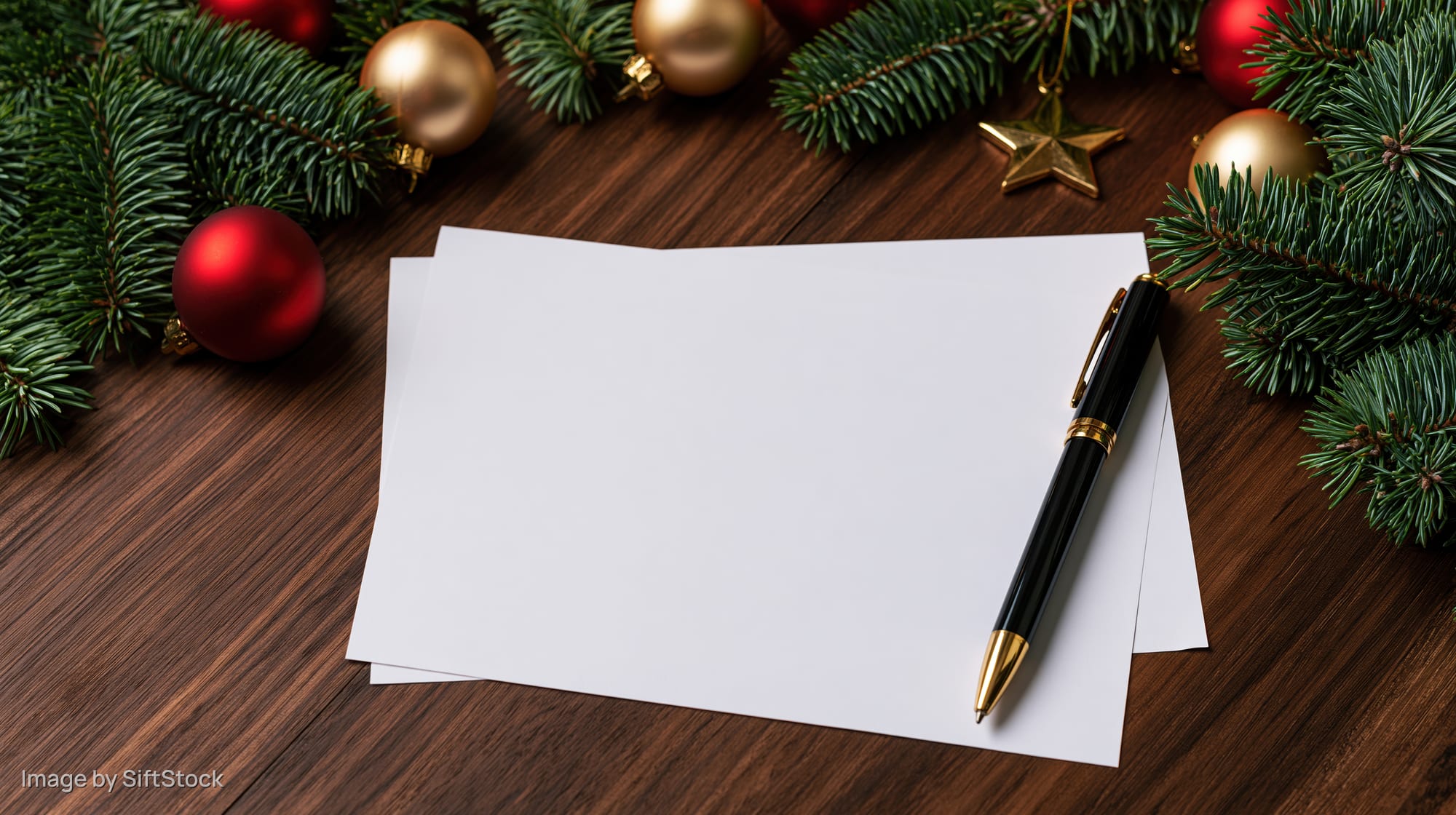 Blank paper and pen on wooden desk surrounded by christmas ornaments and pine branches