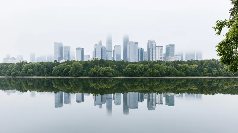 City skyline of glass skyscrapers reflected in calm water behind a dense band of trees