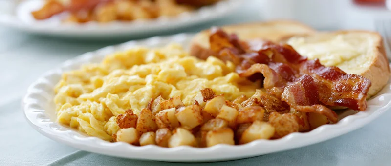 Classic american diner breakfast plate with scrambled eggs bacon potatoes and toast on a white porcelain plate set on a blue cloth tablecloth