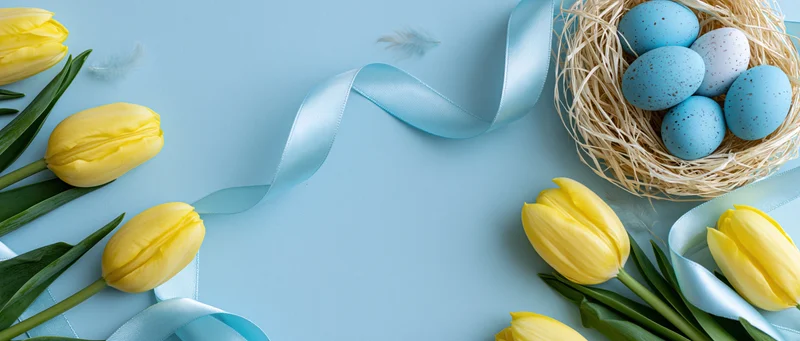 Easter flat lay with yellow tulips, blue speckled eggs in a straw nest, and light blue ribbon on pale blue paper background