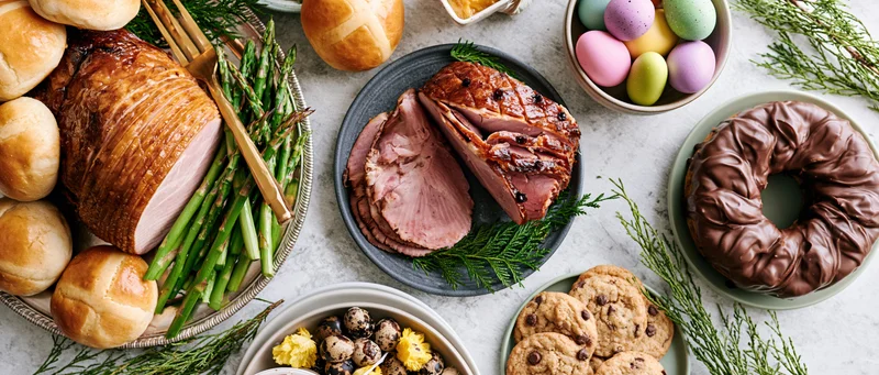 Easter table spread with ham, asparagus, bread, chocolate donut, cookies and pastel eggs arranged overhead on marble surface