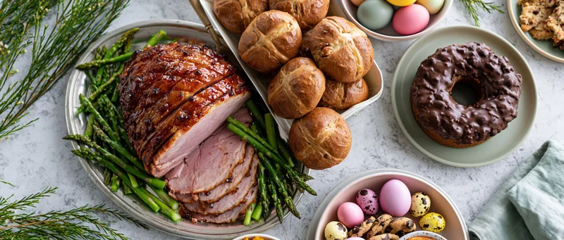Easter table spread with glazed ham, asparagus, bread rolls, chocolate donut, and decorated eggs arranged overhead in flat lay