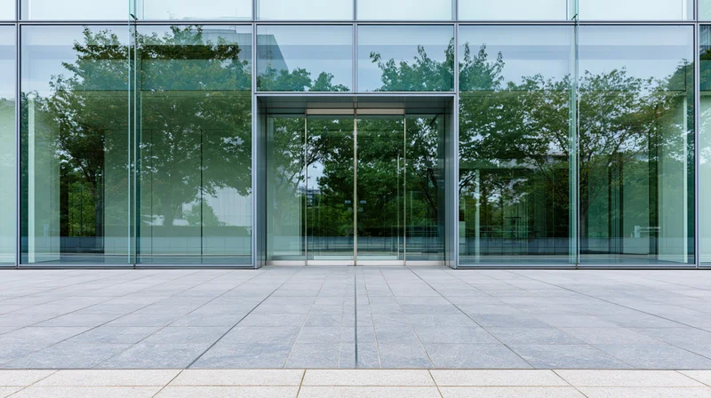 Modern glass office building facade with entrance and tree reflections on tiled plaza