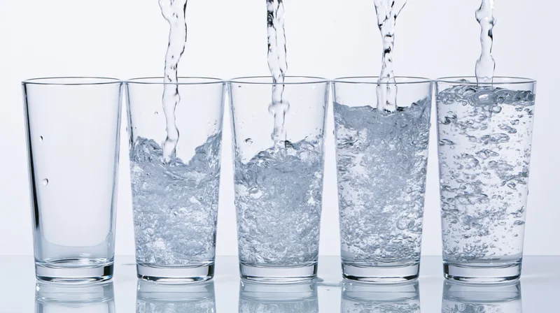 Five tall drinking glasses in a row being filled with water from above on a reflective white surface