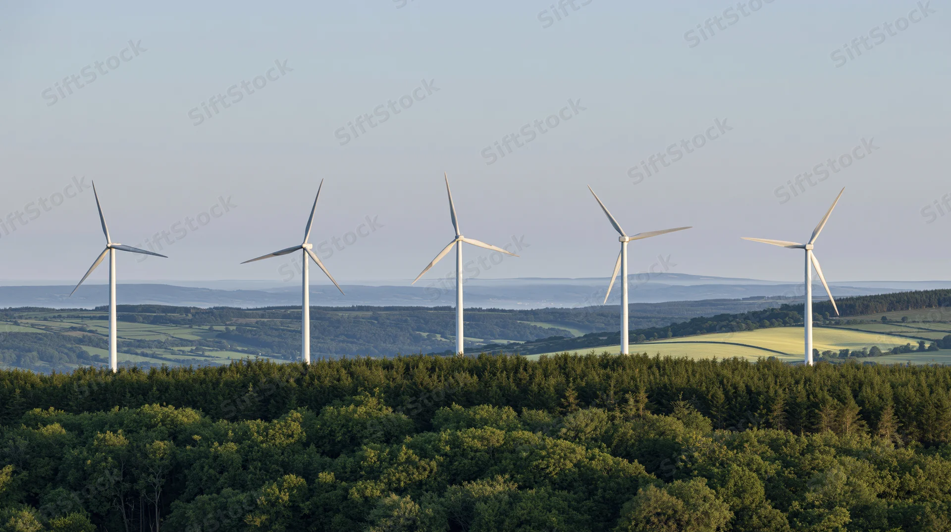 Five white wind turbines on a forested hilltop ridge overlooking a valley