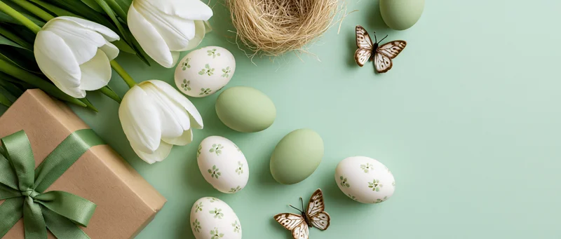 Easter still life flat lay with decorated eggs, white tulips, gift box, butterfly, and nest on mint green background