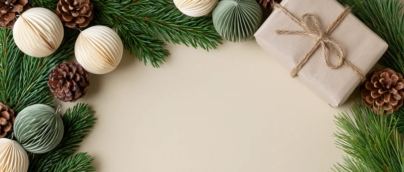 Flat lay of fir branches and pinecones with paper ornaments and wrapped gift forming a border on beige surface