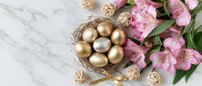Gold and silver painted easter eggs in a nest beside pink alstroemeria flowers on a white marble surface