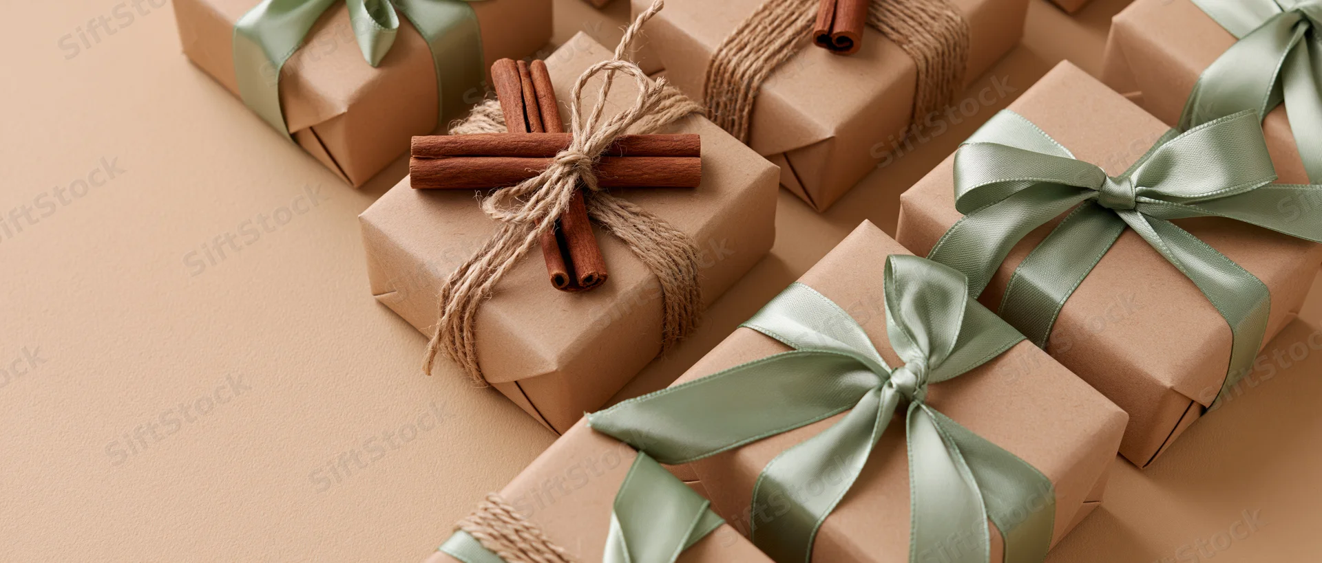 Multiple kraft paper gift boxes wrapped with green satin ribbons and cinnamon sticks arranged in a flat lay on a beige surface