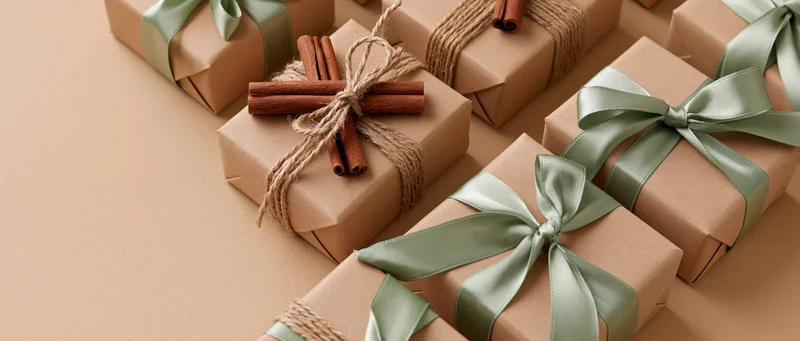 Multiple kraft paper gift boxes wrapped with green satin ribbons and cinnamon sticks arranged in a flat lay on a beige surface