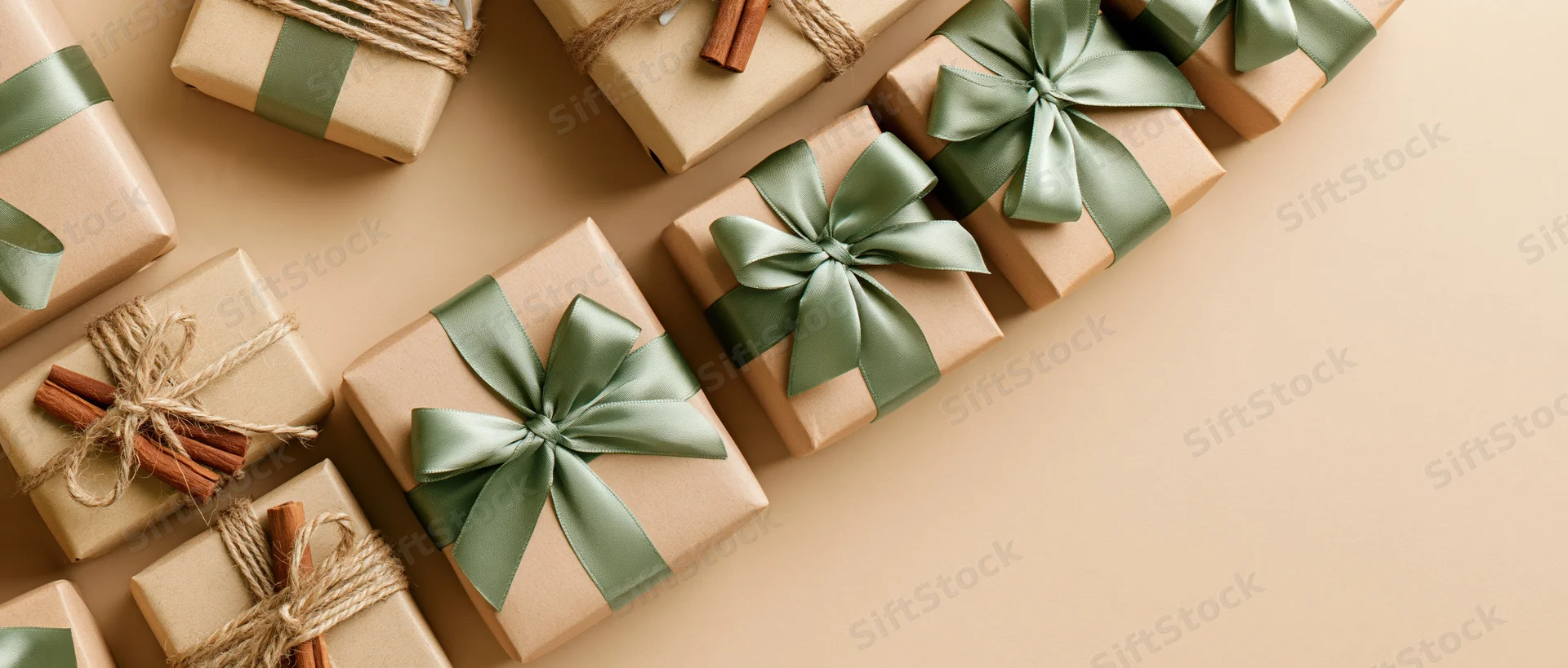 Multiple kraft paper gift boxes with green ribbons and cinnamon sticks arranged in flat lay on beige surface