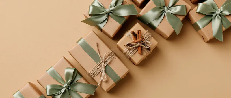 Multiple kraft paper gift boxes with green ribbons and cinnamon sticks arranged diagonally on beige surface