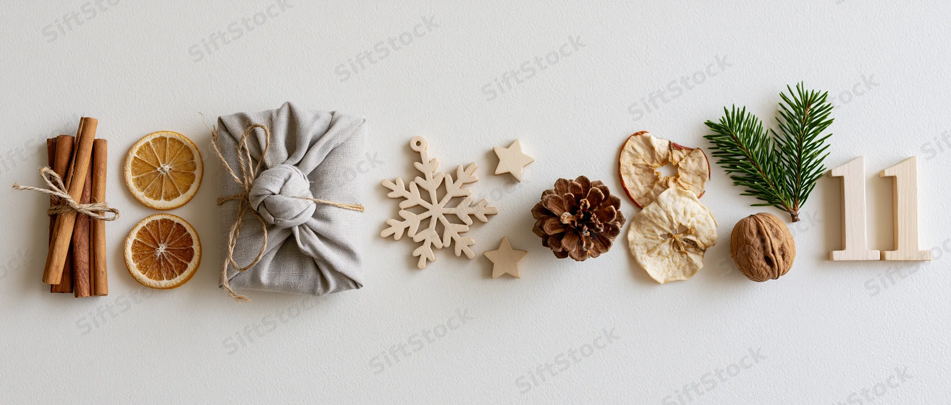 Natural christmas decorations arranged in a horizontal flat lay with cinnamon sticks, dried oranges, a wrapped gift, wooden ornaments, pinecone, walnut, evergreen sprig, and number eleven