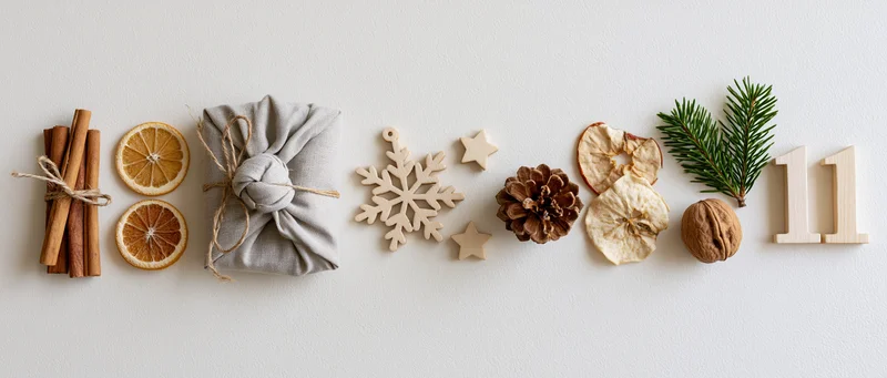 Natural christmas decorations arranged in a horizontal flat lay with cinnamon sticks, dried oranges, a wrapped gift, wooden ornaments, pinecone, walnut, evergreen sprig, and number eleven