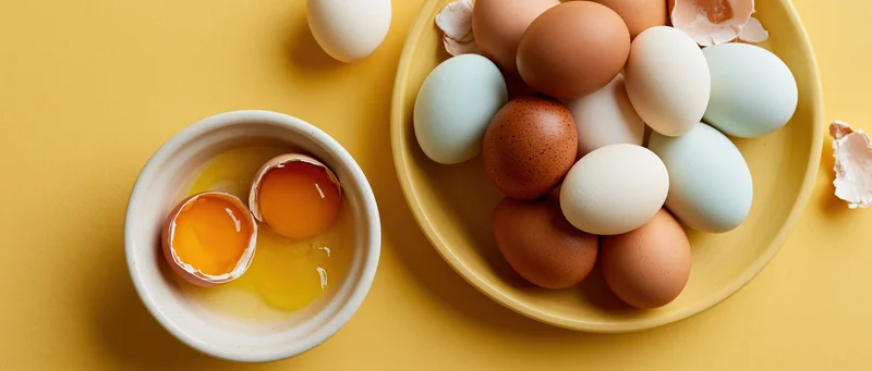 Cracked eggs with orange yolks in a bowl beside a plate of multicolored eggs on yellow surface