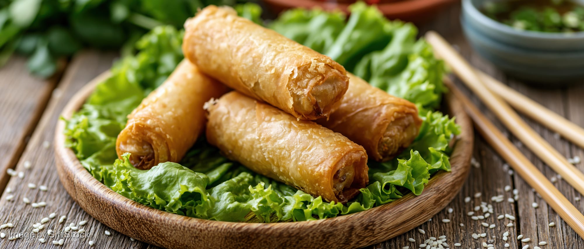 Golden fried spring rolls arranged on a bed of lettuce in a wooden bowl with chopsticks beside on a rustic table