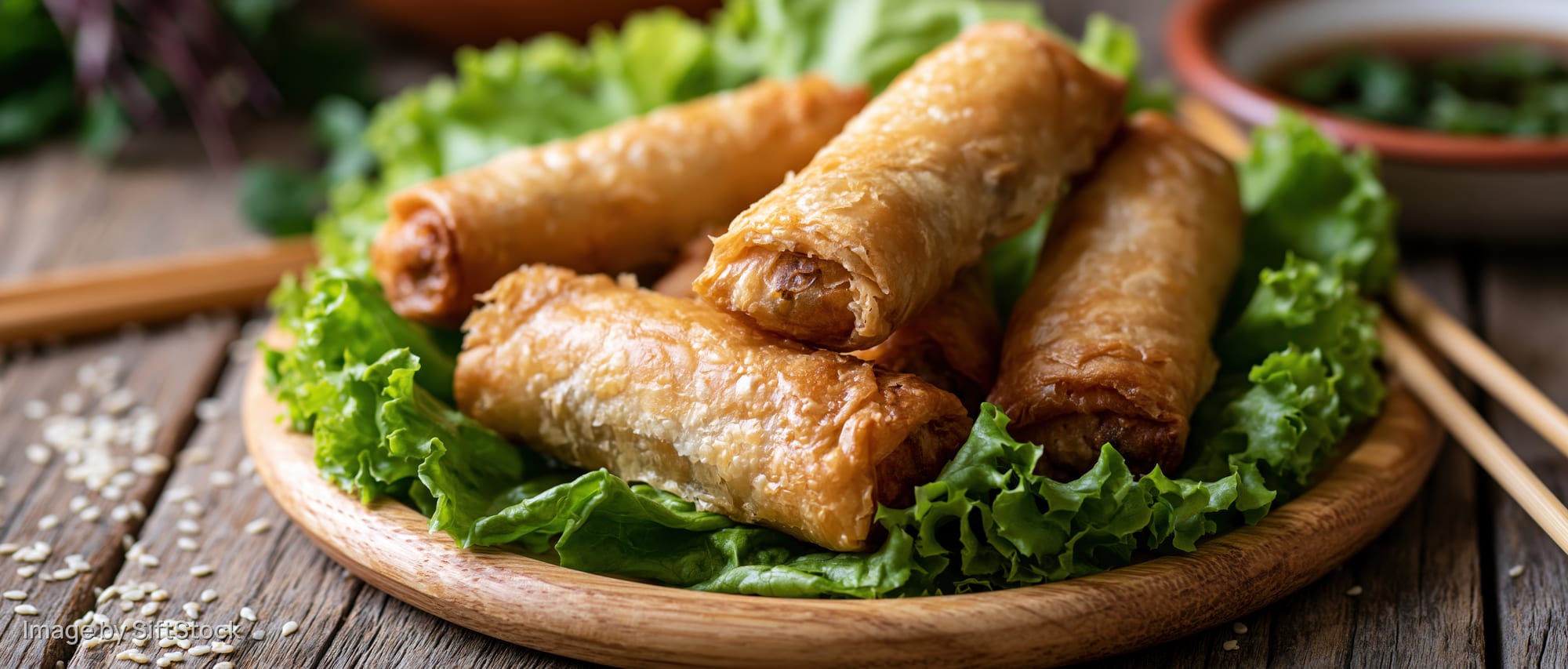 Golden fried spring rolls arranged on a bed of lettuce on a wooden plate with chopsticks nearby