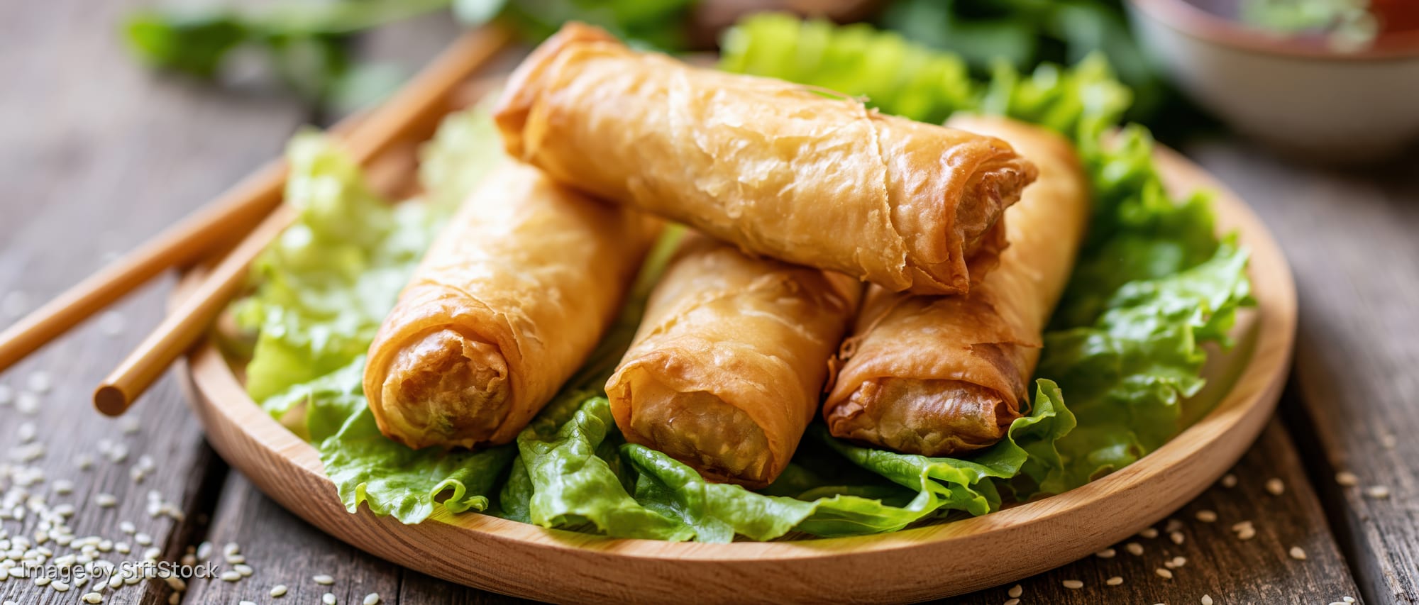 Golden fried spring rolls arranged on a bed of lettuce on a wooden plate with chopsticks nearby