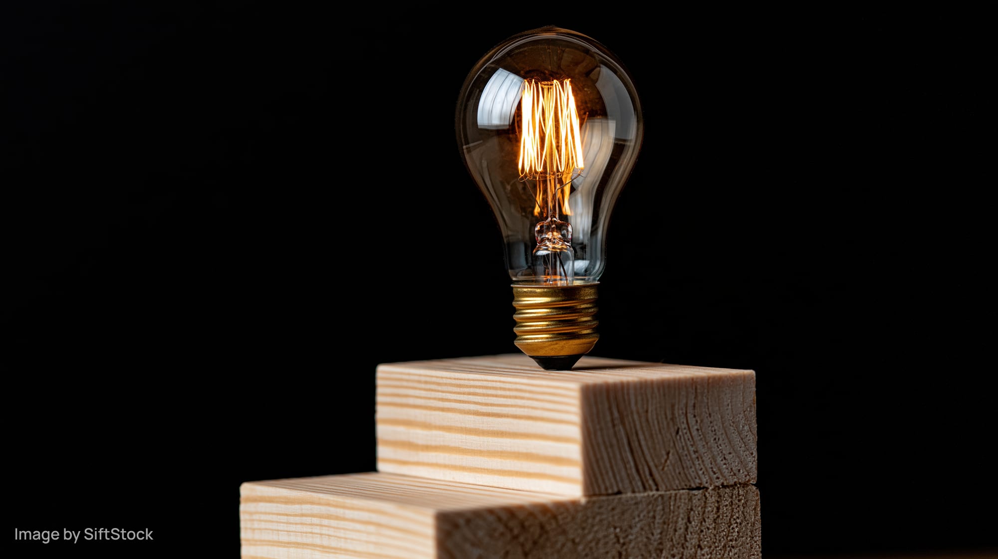 Glowing incandescent light bulb with visible filament resting on stacked wooden blocks against black background