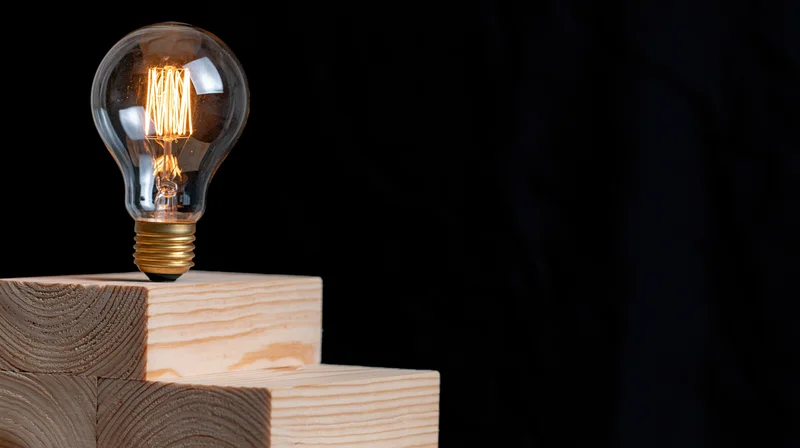 Glowing incandescent light bulb with visible filament resting on stacked wooden blocks against black background