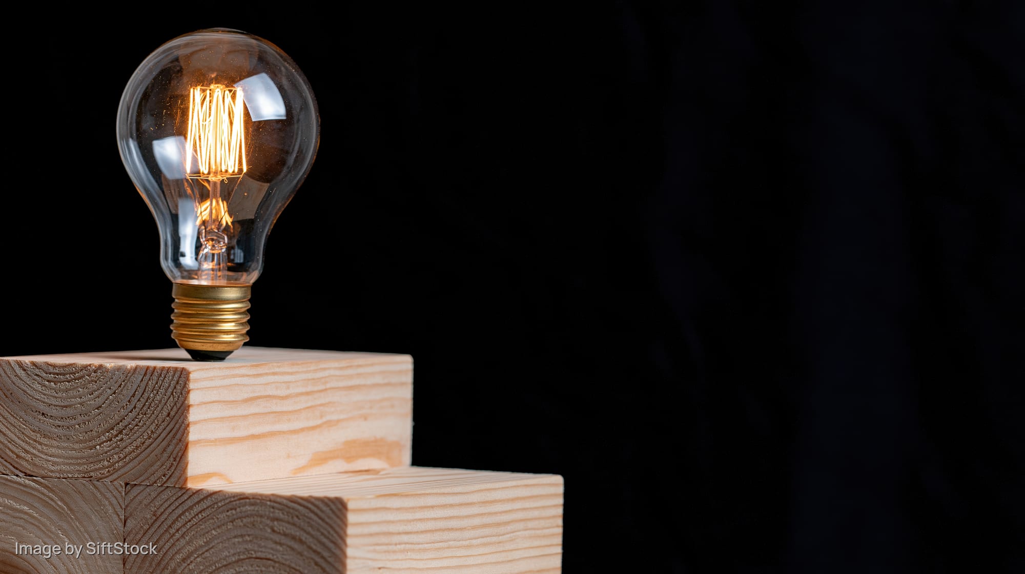 Glowing incandescent light bulb with visible filament resting on stacked wooden blocks against black background