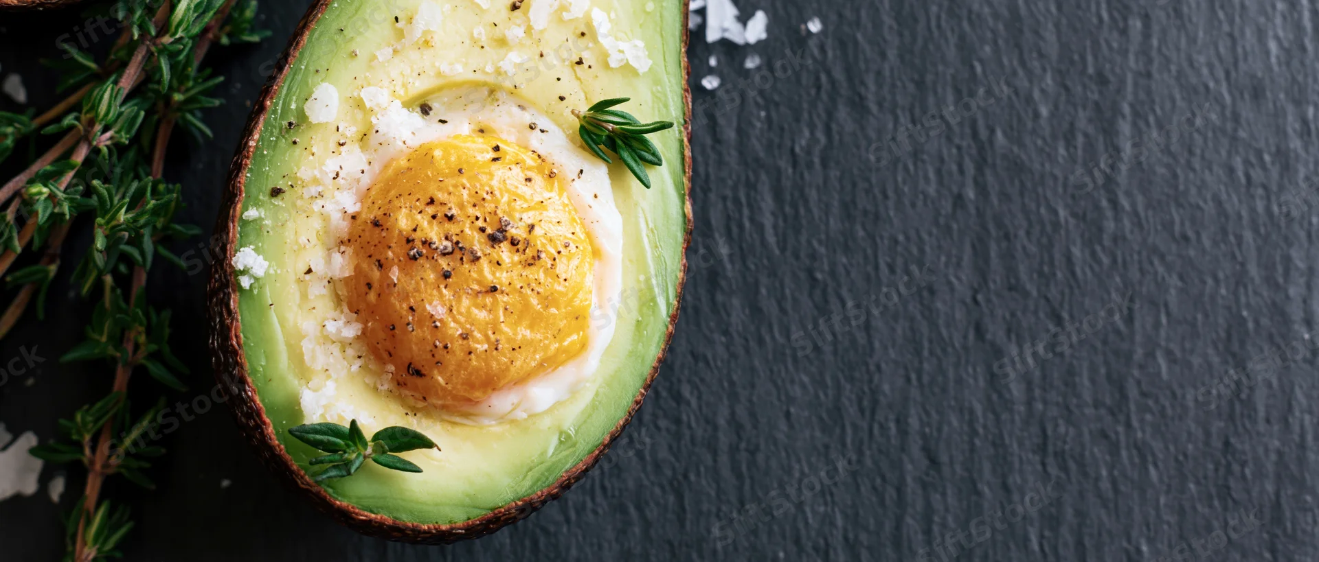 Halved avocado with baked egg nestled in the pit on slate surface garnished with thyme and salt