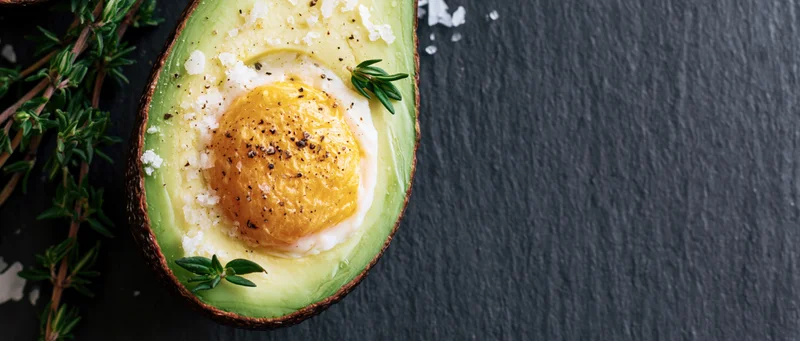 Halved avocado with baked egg nestled in the pit on slate surface garnished with thyme and salt
